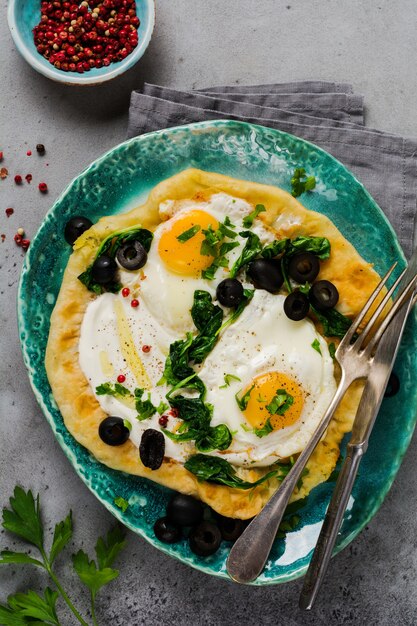 Turkse eieren flatbread met yoghurt, kaas, olijven, spinazie en rode peper op keramische vintage plaat op grijze oude tafel.