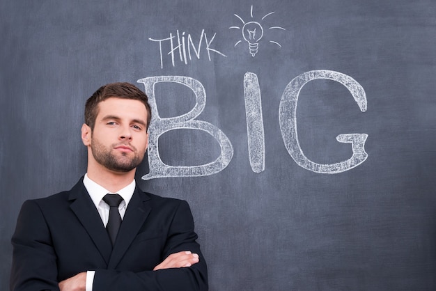 Turn your brains on! Portrait of young businessman keeping arms crossed and looking at camera standing against chalk drawing on the blackboard