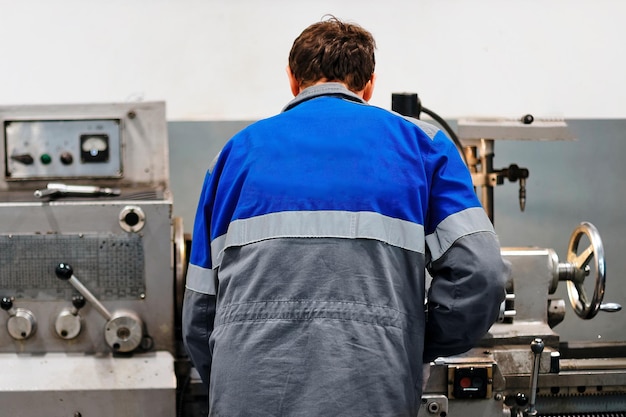 Turner stands behind lathe in production hall and works View of worker from behind from back in overalls