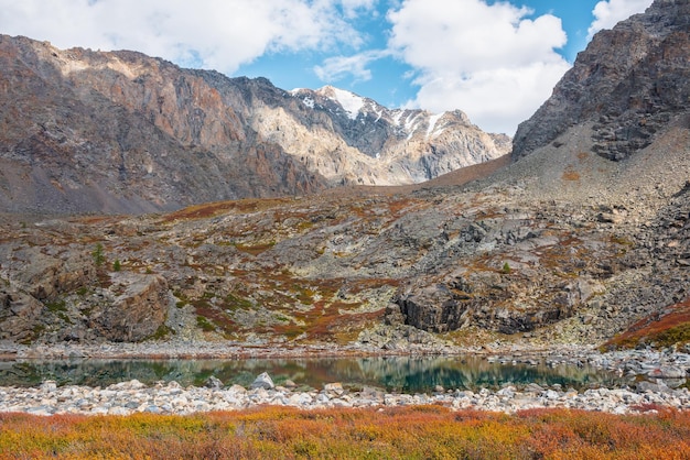 Turquoise bergmeer tussen herfst veelkleurige flora op rotsen tegen hoge rotsachtige bergtop in zonlicht onder wolken in blauwe lucht Zonnelicht alpine meer en grote bergketen Levende herfstkleuren