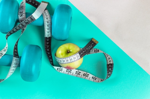 Turquoise dumbbells, tape measure and an apple on a bright turquoise background. 