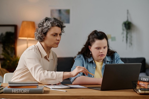 Tutor pointing at monitor of laptop and teaching girl with down syndrome at table at home