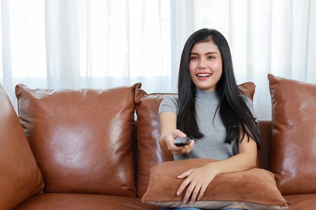 Tv en geluk concept. mooie aziatische vrouw in casual zittend op de bank in de woonkamer, televisie op afstand houdend en kijkend naar iets met een blij lachend gezicht. levensstijlconcept.