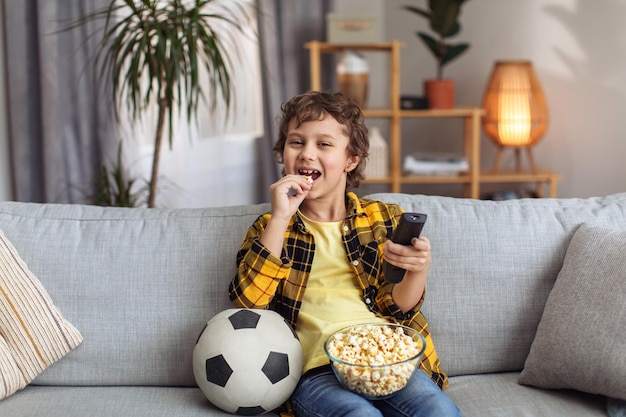 TV 엔터테인먼트 행복한 어린 소년은 집에서 주말을 보내고 스포츠 경기를 보고 팝콘 여유 공간을 먹습니다.