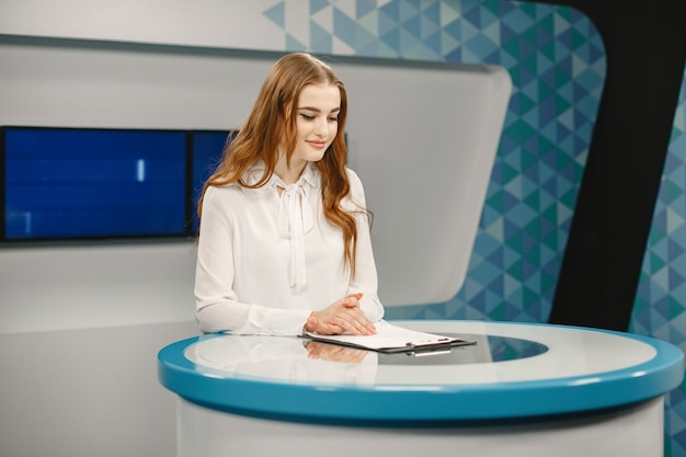 Tv present at studio preparing for new broadcasting. Smiling girl in white shirt sitting at table .