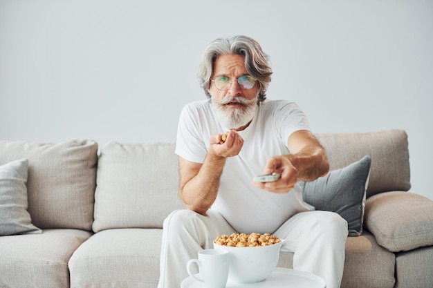 Tv-show kijken en popcorn eten Senior stijlvolle moderne man met grijs haar en baard binnenshuis