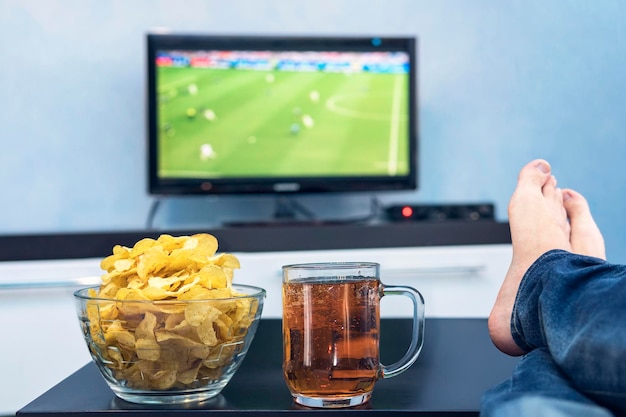 Tv televisie voetbalwedstrijd kijken op tv met hapjes en alcohol relaxen voor de tv Een fan van voetbal kijken speelt een bord chips en een mok bier op tafel