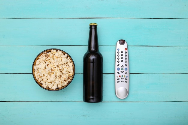 Foto ora della tv. bottiglia di birra, telecomando della tv e ciotola di popcorn su fondo di legno blu. vista dall'alto