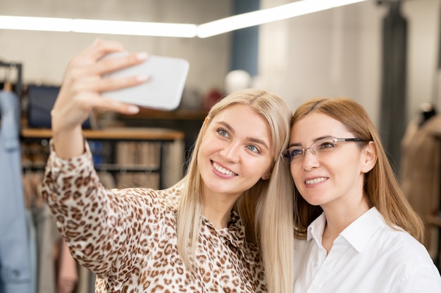 Twee aanhankelijke vrouwelijke consumenten glimlachen tijdens het maken van selfie voor camera op kledingafdeling tijdens het winkelen