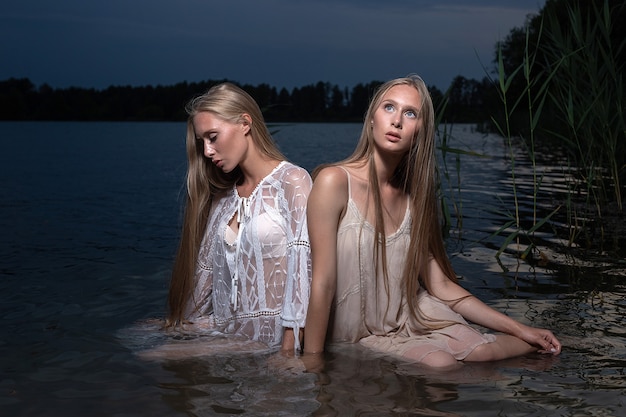 Twee aantrekkelijke jonge tweelingzusjes met lang blond haar poseren in lichte jurken in water van meer op zomeravond
