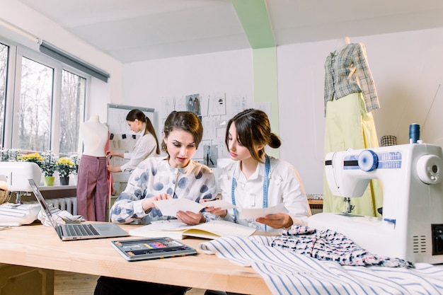 Twee aantrekkelijke vrouwelijke naaistersontwerpers die trendy schetsen en materialen kiezen voor een nieuwe kledingcollectie, terwijl een jonge meisjesontwerper werkt aan een nieuw model, een broek op paspop in de studio aanpassen