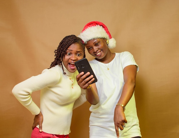 Foto twee afrikaanse dames, zussen of vrienden die vrolijk naar een smartphone kijken terwijl een van hen een kerstmuts op haar hoofd heeft