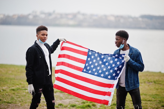 Twee Afrikaanse tienersvrienden met Amerikaanse vlag bij park die medische maskers dragen beschermen tegen infecties en de quarantaine van het coronavirusvirus.