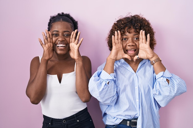 Twee afrikaanse vrouwen die over een roze achtergrond staan en glimlachend vrolijk spelen, gluren een boe-geroep met handen die hun gezicht laten zien, verrast en opgewonden