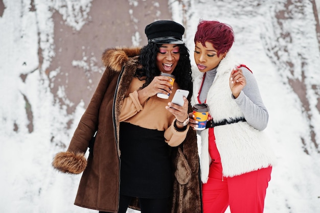 Twee Afro-Amerikaanse dames in schapenvacht en bontjas poseren op winterdag tegen besneeuwde achtergrond met kopjes koffie en kijken naar mobiele telefoon