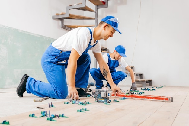 Twee arbeiders in een speciaal uniform leggen tegels met tegelnivelleersysteem en laserwaterpas op de vloer