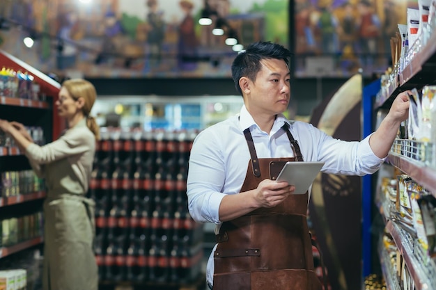 Twee arbeiders in een supermarkt op de kruideniersafdeling regelen producten een aziatische man met een tablet