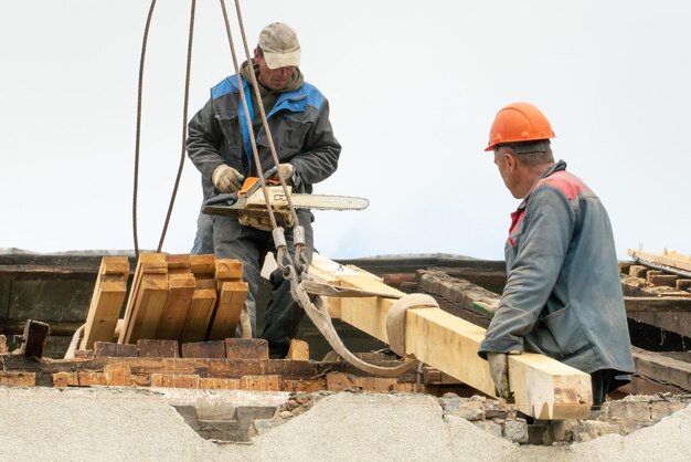 Foto twee arbeiders repareren het dak van een oud gebouw de timmerman houdt een kettingzaag in zijn hand een houten balk is met een touw aan de kraan bevestigd reparatie van een houten dak