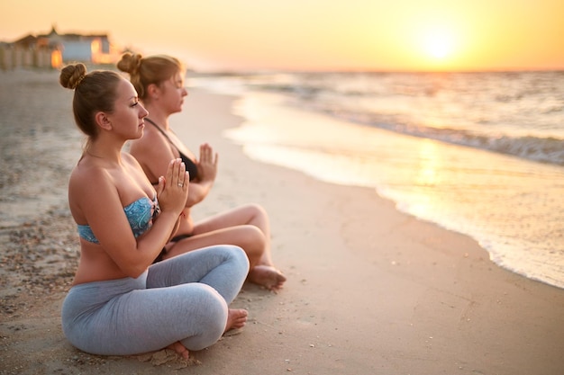 Twee authentieke vrouwen die groepsyoga-meditatie doen op het strand bij zonsopgang, meisjes ontspannen in lotushouding
