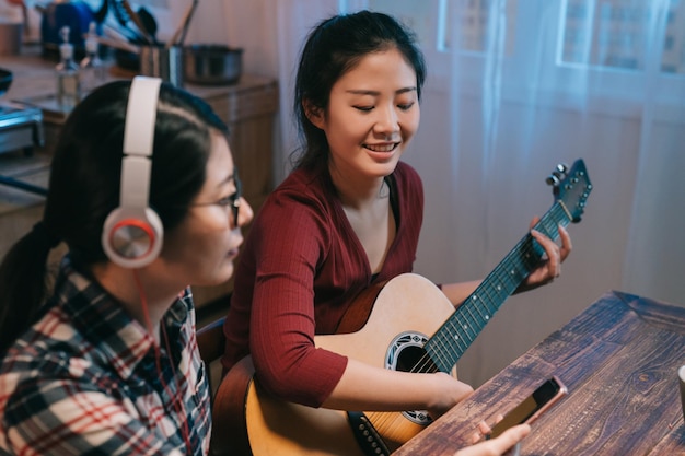 twee Aziatische Chinese vriendinnen ontspannen thuis in de nacht. jonge lachende vrouw gitaar spelen bladmuziek lezen op zus mobiele telefoon. dame met een bril en een koptelefoon zit rustig naast.