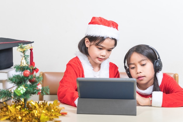 Foto twee aziatische kinderen, kleine meisjes, dragen kerstman die met een vriend aan het chatten is op een digitale tablet. kerstvakantie concept stock photo