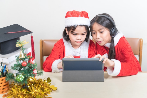 Twee Aziatische kinderen, kleine meisjes, dragen kerstman die met een vriend aan het chatten is op een digitale tablet. Kerstvakantie Concept Stock Photo