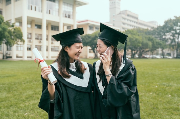 twee aziatische vrouwelijke studenten op afstudeerdag met behulp van slimme telefoon praten. Universitair onderwijs digitale technologie mobiele communicatieconcept. gelukkige meisjes beste vriend op de universiteit lachplezier.