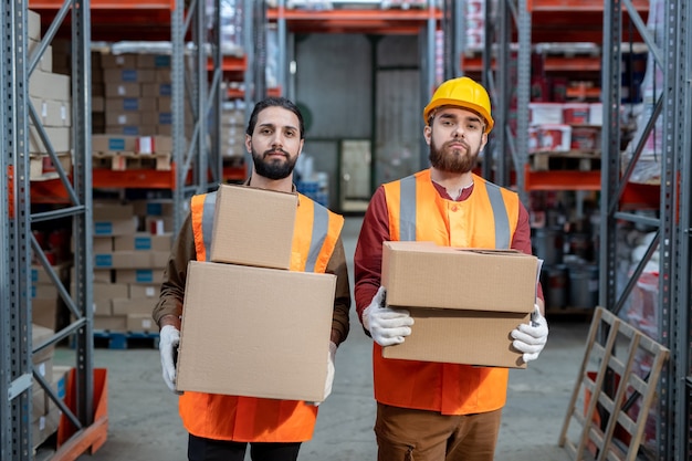 Twee bebaarde werknemers van magazijnen die dozen vasthouden terwijl ze door het gangpad lopen