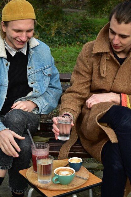 Foto twee blanke mannen, een blonde en een bruinharige, drinken koffie en framboos sap in een park.