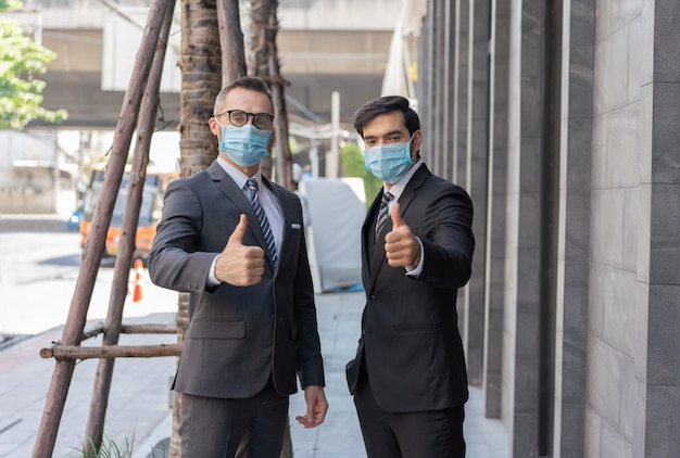 Twee blanke zakenlieden dragen met een medisch masker duimen opdagen tijdens coronavirus covid-19-epidemie op straat