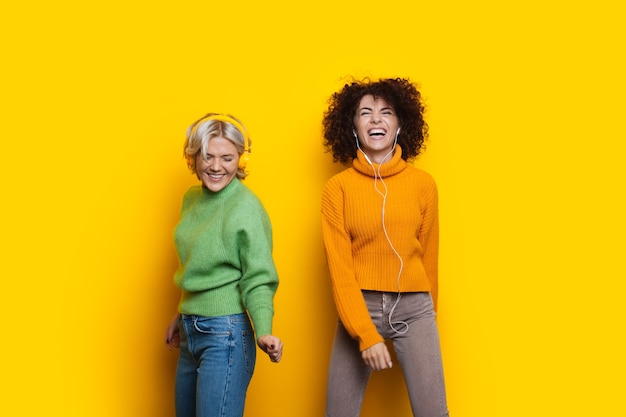 Twee blanke zussen met krullend haar hebben vreugde en dansen op een gele achtergrond met hun koptelefoon