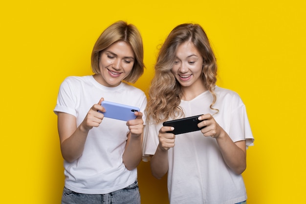 Twee blonde vrouwen met witte t-shirts spelen mobiele games op de telefoon die op een gele muur poseert