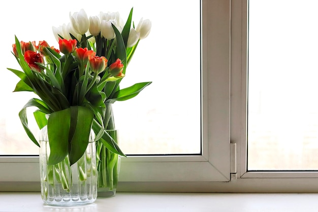 Foto twee boeketten in glazen vazen van oranjerode en witte tulp bloemen op raam tegen de achtergrond van de stad