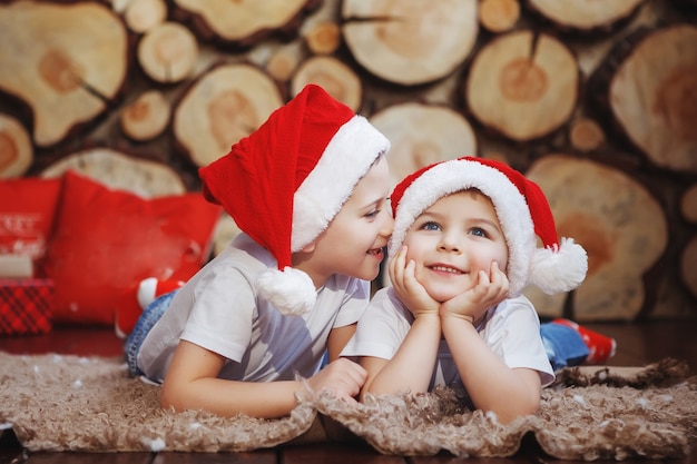 Twee broerjongens met kerstmutsen liggen en lachen op het tapijt in de buurt van de nieuwjaarsboom op de achtergrond van boomsneden.