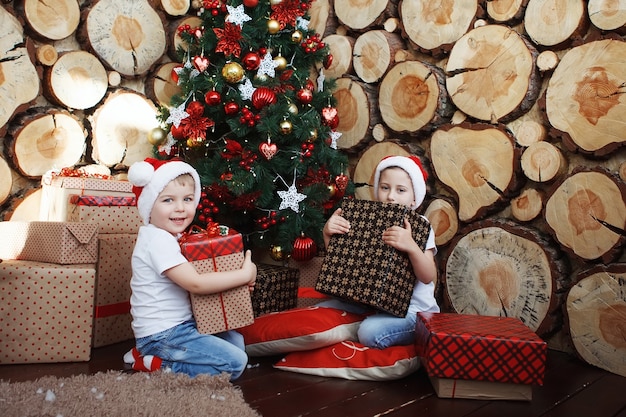 Twee broerjongens met kerstmutsen zitten met geschenkdozen in hun handen op een plaid in de buurt van de nieuwjaarsboom op de achtergrond van boomsneden.