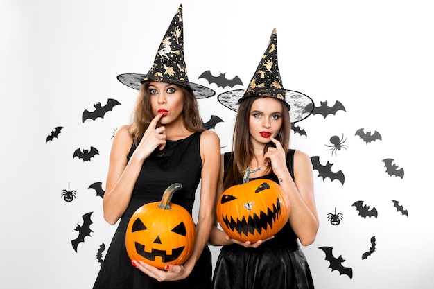 Twee brunette meisjes in zwarte jurken en heksenhoeden houden horror halloween pompoenen op de achtergrond met vleermuizen.