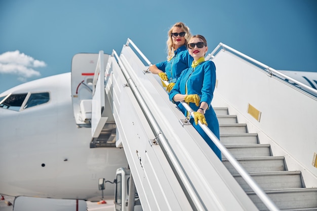 Twee charmante jonge vrouwelijke stewardessen in blauwe pakken die naar de camera kijken en glimlachen terwijl ze op een vliegtuigtrap staan