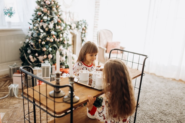 Twee charmante kleine meisjes zitten aan tafel en gaan cacao drinken met Marshmallows en koekjes in de gezellige kamer met de nieuwjaarsboom.