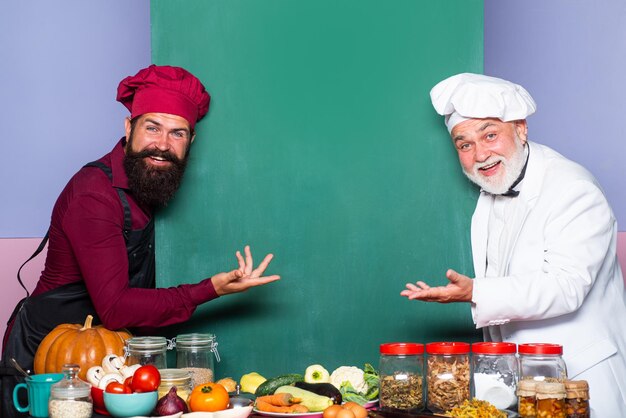 Twee chef-koks in koksmuts wijzende lepel op een leeg bordmenu Gelukkige chef-kok die menubord toont