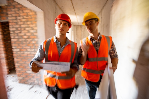 Twee civiel ingenieurs, gekleed in oranje werkvesten en helmen, lopen het gebouw in aanbouw binnen.