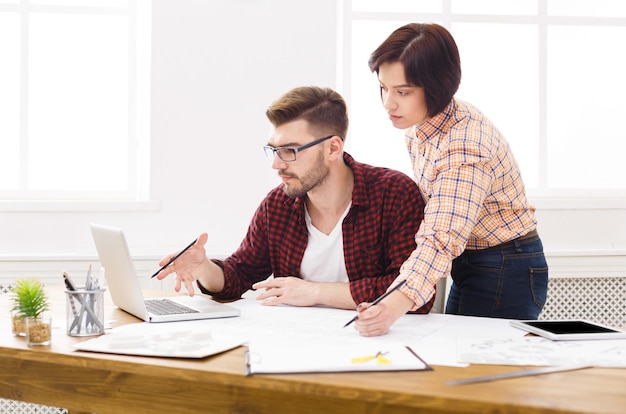 Twee collega's bespreken architectonisch project op laptop, werken met blauwdrukken in modern werkplekinterieur, kopiëren ruimte