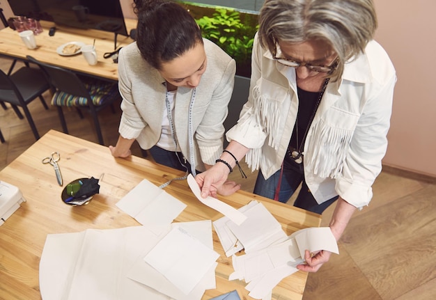 Twee diverse stijlvolle vrouwelijke kleermakers bespreken naaipatronen terwijl ze een nieuwe kledingcollectie ontwikkelen in een modeontwerpworkshop en een kleermakersatelier