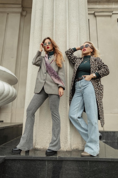 Twee elegante trendy vrouwen vriendinnen met zonnebril in mode herfst kleding met pak jas spijkerbroek en schoenen staat en poseert in de buurt van een vintage kolom in de stad