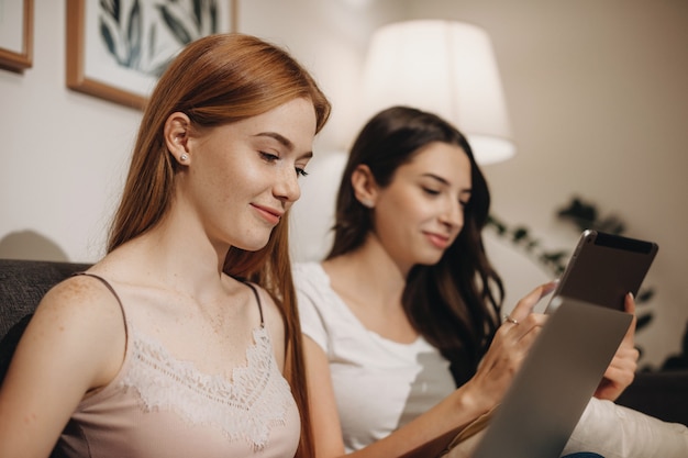 Twee geconcentreerde blanke dames besteden hun tijd zittend op een bank en surfen op internet met behulp van een tablet en laptop