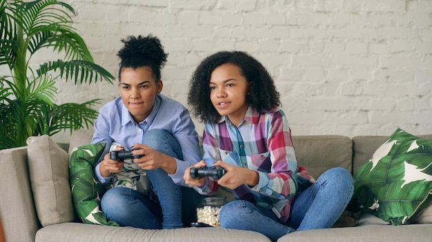 Twee gekrulde meisjes die op de bank zitten, spelen console-computerspellen met gamepad en hebben thuis plezier