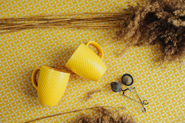 Twee gele thee bekers op tafel. Mesh thee stainer op gele tafel met blad thee rond, theekopjes en gedroogde bloemen op tafel.