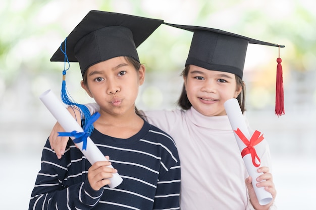 Twee gelukkige afgestudeerde Aziatische vrouwelijke schoolkinderen met een afstudeerpet hebben een opgerold certificaat. Afstuderen Viering Concept Stock Photo