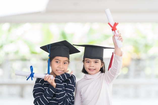 Twee gelukkige afgestudeerde Aziatische vrouwelijke schoolkinderen met een afstudeerpet hebben een opgerold certificaat. Afstuderen Viering Concept Stock Photo