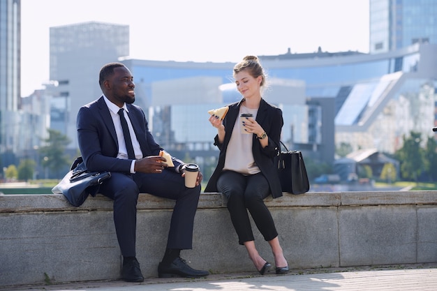 Twee gelukkige jonge interculturele zakenpartners in formele kleding met koffie en lekkere broodjes aan de rivier in stedelijke omgeving