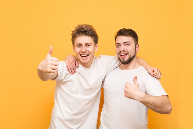 Twee gelukkige jonge mensen die duimen tonen die over geel worden geïsoleerd. twee gelukkige vrienden in witte t-shirts geknuffeld door mannen